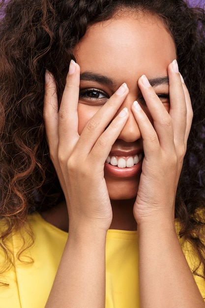 Primer plano de la imagen de la mujer afroamericana positiva con el pelo rizado sonriendo y cubriendo su rostro con las manos aisladas sobre la pared violeta