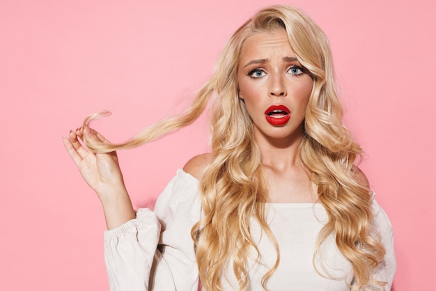 Primer plano de la imagen de una mujer de glamour molesta que expresa su frustración y toca su cabello rubio dañado aislado sobre la pared rosa