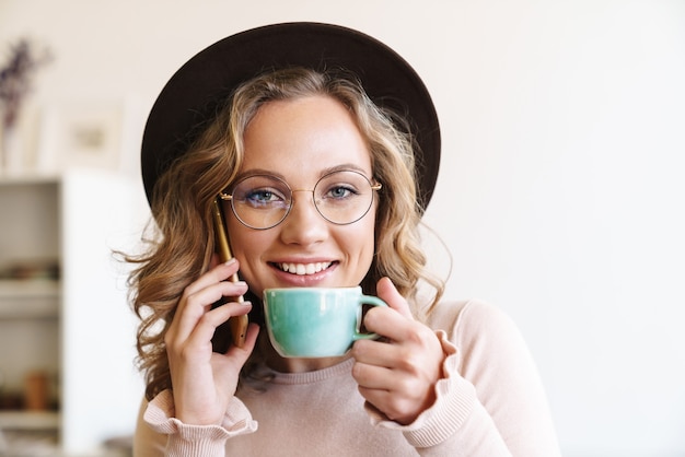 Primer plano de la imagen de una mujer joven alegre en anteojos y sombrero hablando de teléfono móvil y tomando café en casa