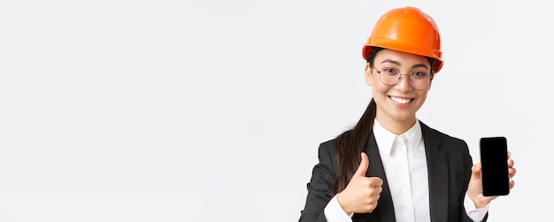 Primer plano de una ingeniera profesional sonriente, gerente de construcción en traje de negocios y casco de seguridad que muestra la pantalla del teléfono inteligente y el pulgar hacia arriba en el fondo blanco de aprobación