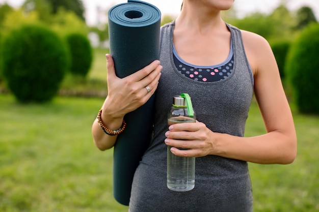 Primer plano de una instructora de yoga con una alfombra de fitness y una botella de agua en una camiseta sin mangas gris.