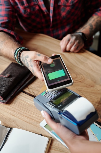 Foto primer plano de irreconocible hombre sentado en la mesa y pagar con tarjeta en línea en el teléfono inteligente en el café