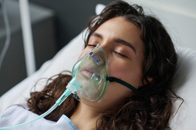 El primer plano de una joven acostada en la cama con los ojos cerrados y respirando con máscara de oxígeno en el hospital