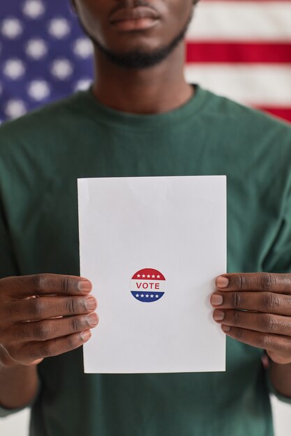 Primer plano de un joven africano que muestra la papeleta durante las elecciones de pie contra la bandera americana