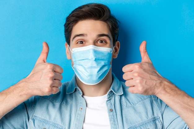 Primer plano de un joven alegre en máscara médica sonriendo, mostrando los pulgares hacia arriba en señal de aprobación, como y de acuerdo, de pie sobre la pared azul