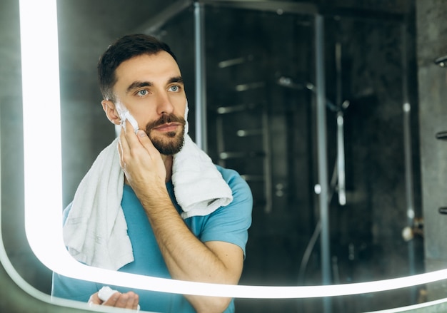 Primer plano de un joven apuesto con una toalla en el cuello de pie frente al espejo y aplicando espuma de afeitar en la cara