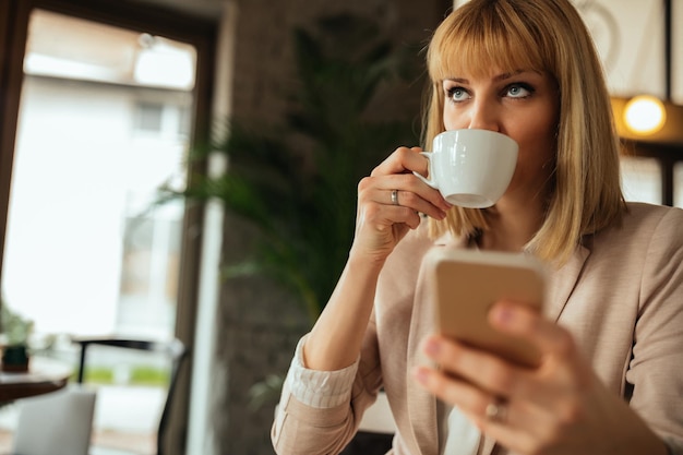 El primer plano de una joven y atractiva mujer de negocios sosteniendo un teléfono móvil mientras bebe café