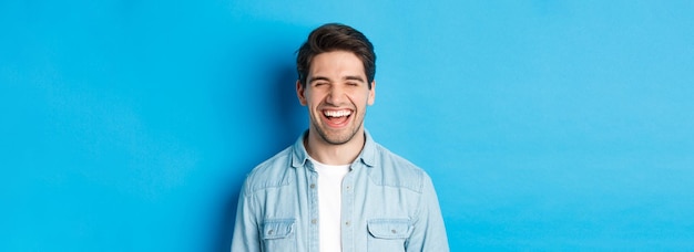 Primer plano de un joven barbudo feliz con ropa casual riendo y sintiéndose alegre de pie sobre el azul