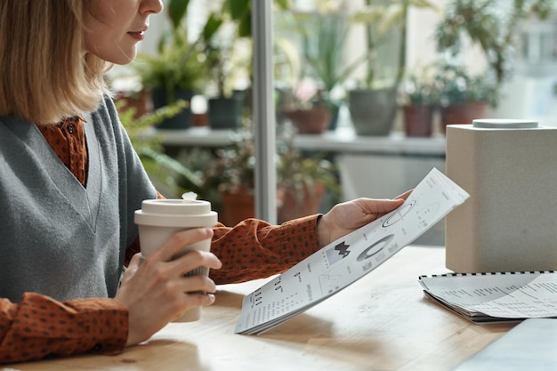 El primer plano de una joven bebiendo café y leyendo un documento con gráficos mientras se sienta a la mesa en