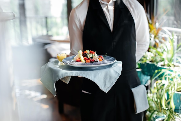 Primer plano de un joven camarero con un elegante uniforme que lleva una exquisita ensalada a un cliente en un hermoso restaurante gourmet Servicio de mesa en el restaurante