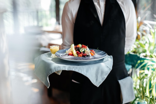 Primer plano de un joven camarero con un elegante uniforme que lleva una exquisita ensalada a un cliente en un hermoso restaurante gourmet. Servicio de mesa en el restaurante.