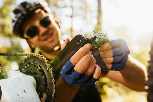 Primer plano de un joven ciclista concentrado y sonriente con gafas de casco y guantes sentado frente a su bicicleta de pedales de reparación de bicicletas antes de hacer ejercicio en bicicleta al aire libre en el bosque