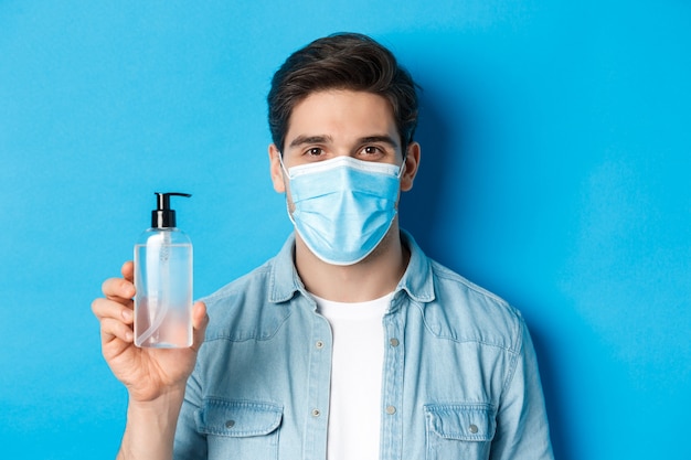 Primer plano de un joven en el consejo de la máscara médica para usar desinfectante de manos, mostrando una botella antiséptica, de pie sobre una pared azul