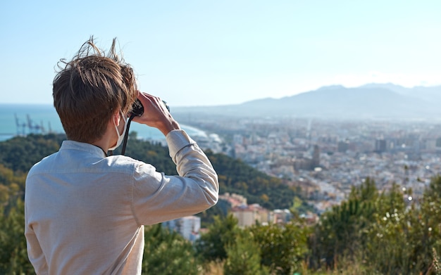 Un primer plano de un joven de España con una camisa blanca con una máscara observando la ciudad costera a través de binoculares