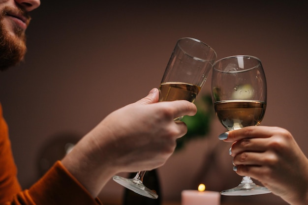 El primer plano de un joven feliz irreconocible que tintinea copas de vino con su esposa en la mesa con velas en una habitación oscura