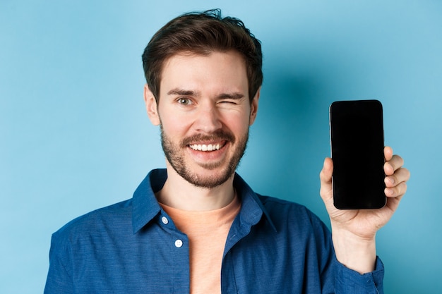 Primer plano de un joven guapo sonriendo, guiñando un ojo y mostrando la pantalla del teléfono móvil vacía, de pie en ropa casual sobre fondo azul.