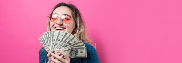 El primer plano de una joven hermosa mujer con un suéter azul en gafas con dinero en dólares americanos en la mano sobre fondo rosa Expresa emociones y gestos de alegría