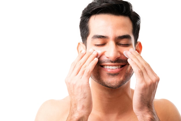 Foto primer plano de un joven hispano aplicando crema humectante en las mejillas con los dedos