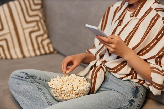 Primer plano de una joven irreconocible relajándose en casa con su teléfono inteligente y comiendo palomitas de maíz.