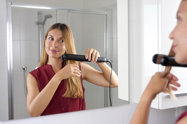 Primer plano de una joven latina usando plancha de vapor para peinar el cabello en el espejo del baño