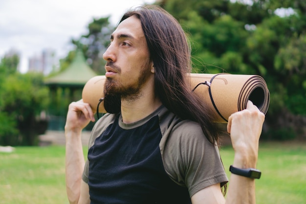 Primer plano de un joven latino e hispano con barba y cabello largo usando un reloj inteligente que lleva la estera de yoga sobre sus hombros en un campo verde