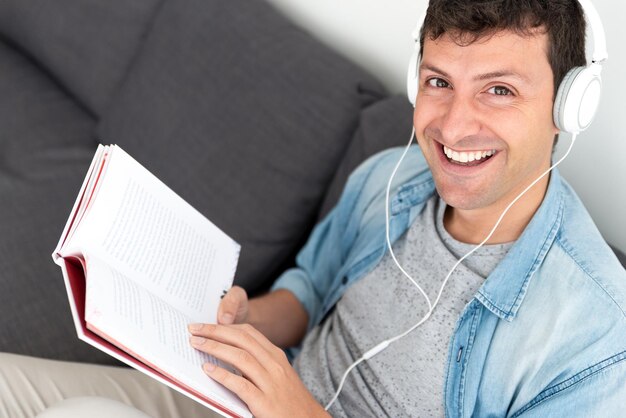 Primer plano de un joven mirando a la cámara mientras lee un libro y escucha música con auriculares