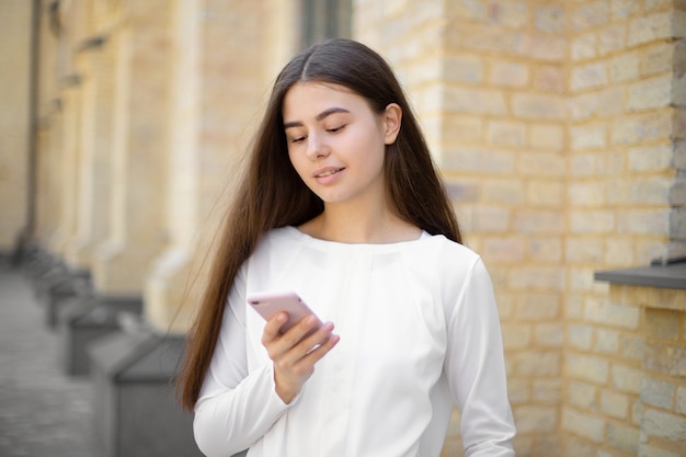 Foto primer plano de una joven morena con un pelo largo en blanco usando el teléfono cuando camina por la calle