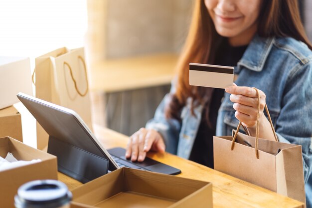 Primer plano de una joven mujer asiática con tablet pc y tarjeta de crédito para compras en línea con paquete postal postal y bolsas de compras sobre la mesa