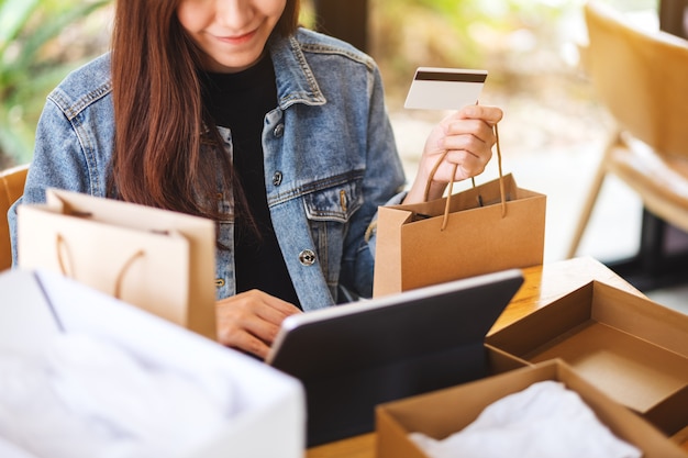 Primer plano de una joven mujer asiática con tablet pc y tarjeta de crédito para compras en línea con paquete postal postal y bolsas de compras sobre la mesa