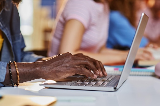 Primer plano de un joven negro escribiendo en el teclado de una computadora portátil mientras estudiaba en la biblioteca universitaria con un estudiante