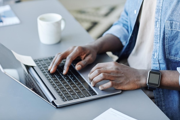 Primer plano de un joven negro escribiendo en el teclado de una computadora portátil mientras trabaja de forma remota desde el espacio de copia en casa