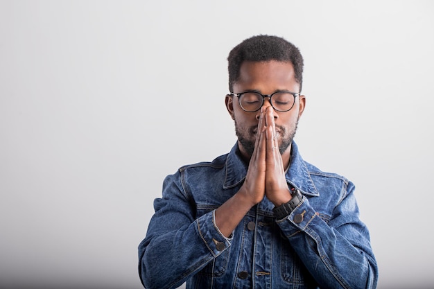 Primer plano de un joven negro con gafas orando con las palmas juntas y los ojos cerrados, pidiendo al Dios buena suerte salud de éxito sobre fondo gris con espacio de copia. La emoción humana, el concepto de creencia