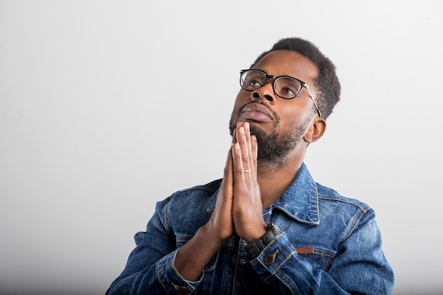 Primer plano de un joven negro rezando con las manos juntas, pidiendo a Dios buena suerte, éxito, salud, mirando hacia arriba, pared gris aislada del estudio. La emoción humana, la expresión facial, el concepto de creencia.