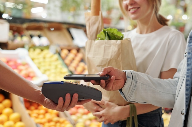 Foto primer plano de la joven pareja pagando productos frescos con smartphone en el mercado de alimentos orgánicos