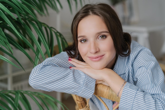 Primer plano de una joven de pelo bastante oscuro con una sonrisa encantadora y gentil que se apoya en las manos posa en el interior cerca de una plantación verde con manicura roja maquillaje Concepto de ocio y relajación de la gente