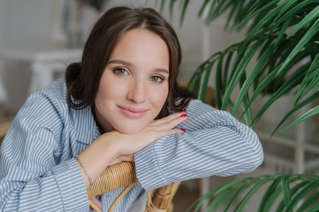 Primer plano de una joven de pelo bastante oscuro con una sonrisa encantadora y gentil que se apoya en las manos posa en el interior cerca de una plantación verde con manicura roja maquillaje Concepto de ocio y relajación de la gente