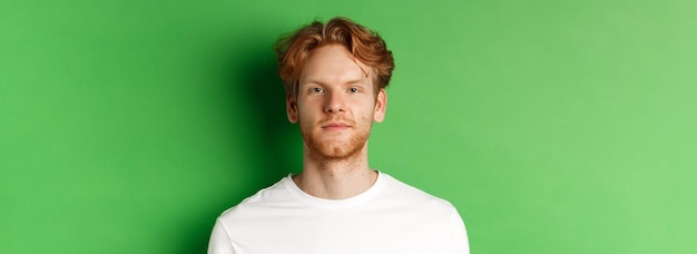 Primer plano de un joven con el pelo rojo y la barba desordenada mirando a la cámara de pie sobre fondo verde