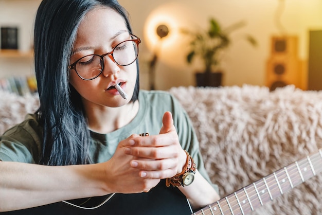 Primer plano de una joven que enciende un porro de marihuana mientras sostiene la guitarra en casa