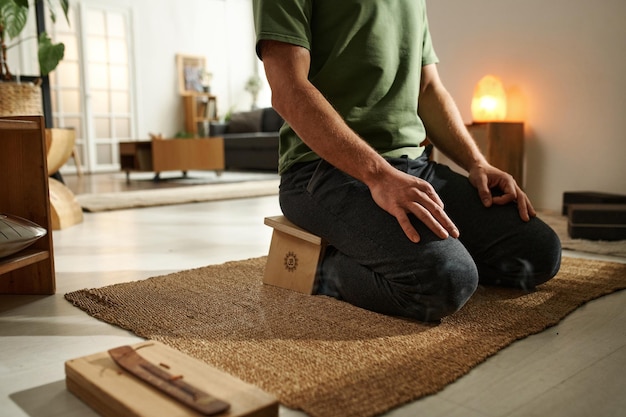 El primer plano de un joven sentado en la alfombra de la habitación y meditando con un palo de aroma