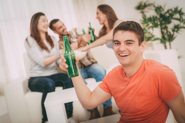 Primer plano de un joven sonriente en la fiesta en casa con cerveza y vítores. Sus amigos en el fondo. Enfoque selectivo. Centrarse en primer plano.