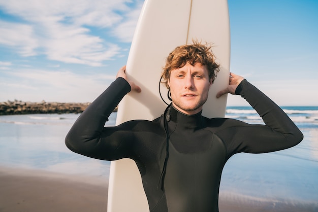 Primer plano de joven surfista de pie en la playa con su tabla de surf y vistiendo traje de surf negro. Concepto de deporte y deporte acuático.
