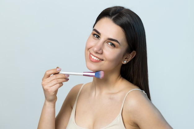 Primer plano de una joven en un top claro sobre un fondo blanco haciendo un maquillaje facial. Una mujer bonita sostiene un cepillo cosmético cerca de su rostro y sonríe.