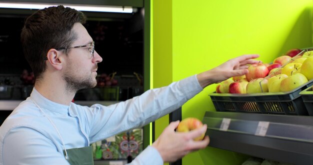 Primer plano de un joven trabajador con delantal verde arreglando manzanas en el supermercado Personas y profesión Tienda de abarrotes Consumismo