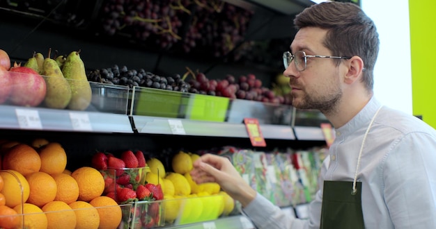 Primer plano de un joven trabajador con delantal verde que almacena los limones en el supermercado Joven empleado en el trabajo Hombre guapo arreglando frutas en el estante Gente y profesión Tienda de abarrotes Consumismo