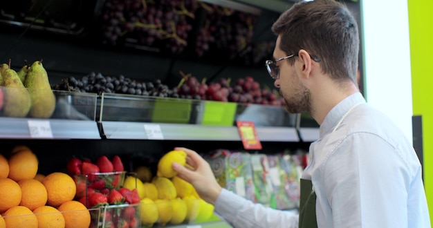Primer plano de un joven trabajador con delantal verde que almacena los limones en el supermercado Joven empleado en el trabajo Hombre guapo arreglando frutas en el estante Gente y profesión Tienda de abarrotes Consumismo