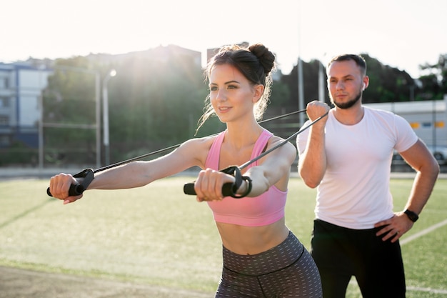 Foto primer plano de jóvenes deportistas entrenando en el estadio en el verano, una mujer haciendo ejercicios con un expansor y un hombre ayudándola