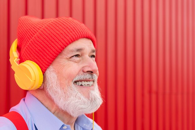 Primer plano de un jubilado sonriente divertido que usa auriculares para escuchar música aislada en un fondo rojo mirando hacia otro lado