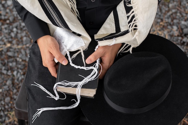 Primer plano de un judío sosteniendo un siddur sobre el que descansa el tzitzit de su talit y junto a él un sombrero