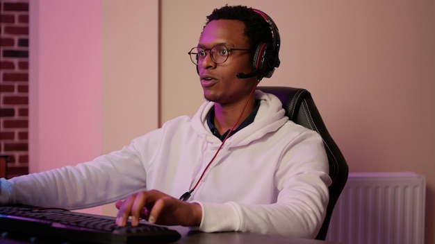 Primer plano de un jugador afroamericano con auriculares para juegos hablando con miembros del equipo en un juego multijugador en pc profesional. Hombre mirando la pantalla de la computadora mientras juega y transmite la competencia en línea.