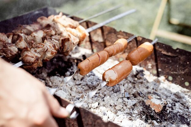 Un primer plano de kebabs en broches, un hombre asando carne al aire libre.